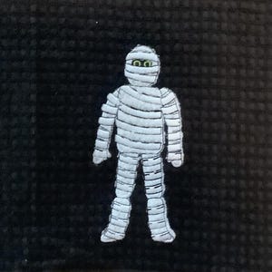 May include: A white mummy figure with green eyes, wrapped in bandages, stands against a black background. The mummy is a Halloween-themed decoration, with detailed stitching and a cartoonish design.