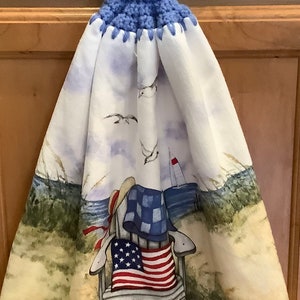 May include: A white kitchen towel with a beach scene featuring a beach chair with an American flag, seagulls, and a sailboat. The towel has a blue crocheted loop with a white button.