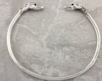May include: A silver bangle bracelet with two dog heads on the ends. The dog heads are facing each other.