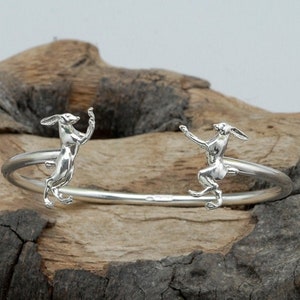 May include: A silver bangle bracelet with two sculpted hare figures on either side. The hares are facing each other and appear to be leaping.