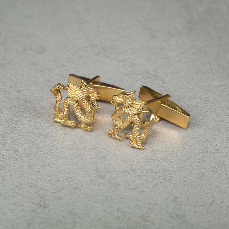 May include: A pair of gold-tone cufflinks featuring a detailed dragon design. The cufflinks are connected by a simple bar.