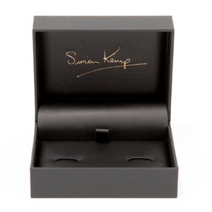 May include: A black rectangular box with a hinged lid. The inside of the box is lined with black velvet and has two circular indentations. The lid has the inscription "Simon Kemp" in gold lettering.