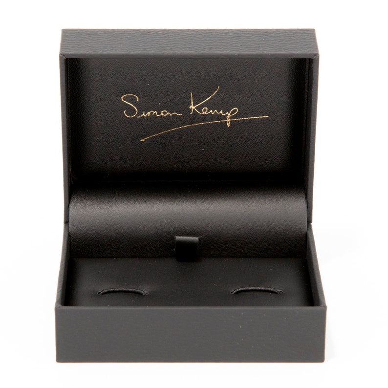 May include: A black rectangular box with a hinged lid. The inside of the box is lined with black velvet and has two circular indentations. The lid has the inscription "Simon Kemp" in gold lettering.