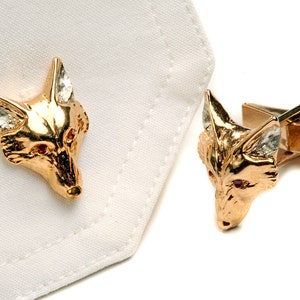 May include: A pair of gold fox head cufflinks with red gemstone eyes. The cufflinks are on a white background.