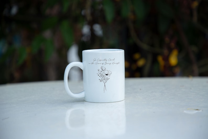 May include: White ceramic mug with a curved handle. The mug features a black floral design and the text "So Casually Chic in the Name of Being Honest". The mug is sitting on a white surface.