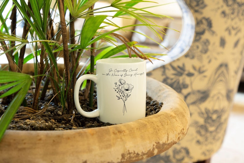 May include: White ceramic mug with a black floral design and the text "So Casually Cruel in the Name of Being Honest." The mug is in a large, weathered planter with green plants.
