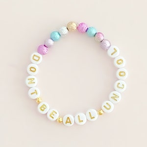 Puede incluir: Pulsera de cuentas blancas con letras doradas que deletrean "Don't Be All Uncool". Las cuentas están intercaladas con cuentas iridiscentes de color rosa, azul y morado.