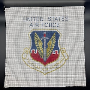 May include: A square, off-white canvas with the words "UNITED STATES AIR FORCE" at the top. The center features a shield emblem with a sword, wings, and the Air Force logo in blue, red, and gold.