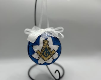 Peut inclure: Ornement décoratif bleu et blanc avec un symbole maçonnique. L'ornement rond présente un motif en forme d'étoile blanche avec un symbole doré et bleu au centre. Il est suspendu à un support en métal noir avec un ruban blanc.