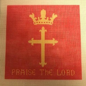 May include: A needlepoint canvas with a red background featuring a gold crown and cross design. The words "PRAISE THE LORD" are stitched in gold at the bottom. The canvas is bordered by a white mesh.