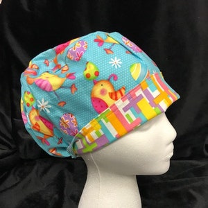 May include: A blue scrub hat with a colorful pattern of birds, eggs, and flowers. The hat has a bright, multi-colored trim.