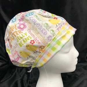 May include: A white scrub hat with a colorful Easter-themed print. The print features text that says "Happy Easter", "Jelly Beans", "Colored Eggs", and "Easter Bunnies".