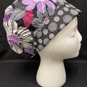 May include: A black scrub hat with a floral print featuring purple, pink, and white flowers on a black background. The hat has a contrasting black and white polka dot pattern on the inside.