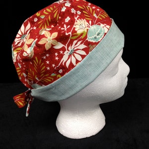 May include: A red and teal patterned scrub hat with a tie. The hat has a floral design with white flowers and green leaves.