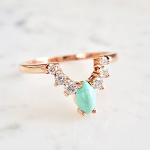 May include: Rose gold ring with a turquoise teardrop-shaped stone at the centre, encircled by a curved row of small, clear gemstones. The ring's band is thin and delicate, with a polished finish. The ring is set against a white marble surface.