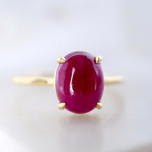 May include: A gold ring featuring a large, oval-shaped ruby gemstone. The ruby is a deep red colour and has a smooth, polished surface.