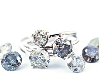 Puede incluir: Un conjunto de anillos de plata con diamantes grises y negros. Los anillos están apilados uno encima del otro y hay varias piedras sueltas esparcidas a su alrededor.