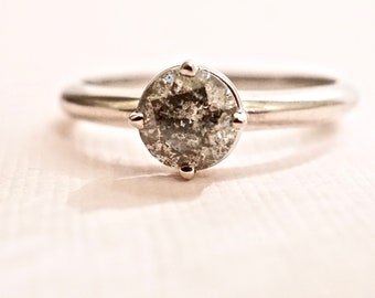 Puede incluir: Un anillo de tono plateado con un diamante redondo de tipo sal y pimienta en un engaste de cuatro garras. La banda es lisa y pulida, mostrando un brillo metálico que complementa la apariencia moteada, gris y blanca de la piedra central.