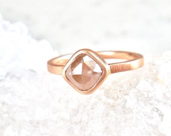 Op de afbeelding: Een roségouden ring met een vierkante, lichtbruine edelsteen. De edelsteen is gezet in een bezel-zetting en de band heeft een eenvoudig, gepolijst ontwerp. De ring wordt gepresenteerd op een wit, getextureerd oppervlak.