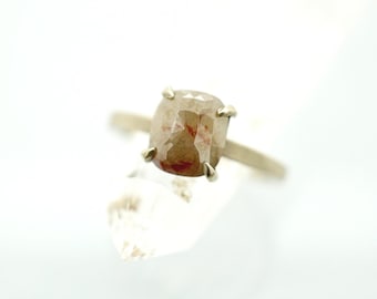 Op de afbeelding: Een gouden ring met een grote, kussenvormige, lichtbruine diamant met roodachtige insluitsels. De diamant wordt vastgehouden door vier klauwen. De ring staat tegen een witte achtergrond, waardoor de kleur en helderheid van de edelsteen worden benadrukt.