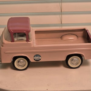 May include: A pink toy pickup truck with a white cab and a brown bed. The truck has a white steering wheel, black tires, and a sticker that says "No. 6200 NYLINT KENNELS".