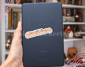 May include: A black Kindle e-reader with the word "Kindle" printed on the device. A sticker with the words "Fluent in Literature" is placed on the device. The background includes a bookshelf.