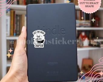 May include: A black Kindle e-reader with a white sticker showing a skeleton reading a book, with the text "Just One More Chapter". The e-reader is held in a hand, with a bookshelf visible in the background. The sticker is die cut.