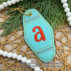 May include: A turquoise keychain with the letter "A" embroidered in orange. The keychain has a gold-colored clasp and is outlined with yellow stitching. The design includes two horizontal maroon stripes. The Etsy shop URLs are visible at the bottom.