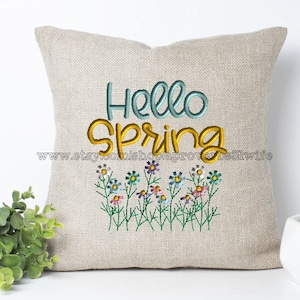 Può includere: Un cuscino beige con testo ricamato che recita "Hello Spring" in blu e giallo. Il cuscino è decorato con una fila di fiori colorati.