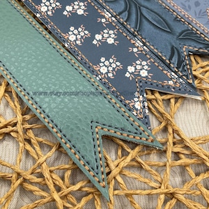 May include: Close-up of several decorative bookmarks with a stitched border. The bookmarks feature various patterns, including floral and textured designs, in shades of green, blue, and white. The background is a woven texture.