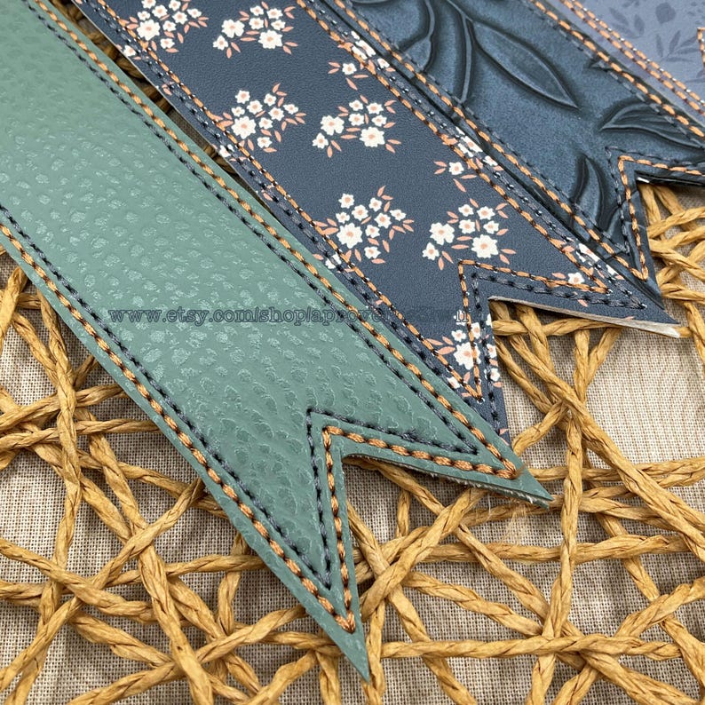 May include: Close-up of several decorative bookmarks with a stitched border. The bookmarks feature various patterns, including floral and textured designs, in shades of green, blue, and white. The background is a woven texture.