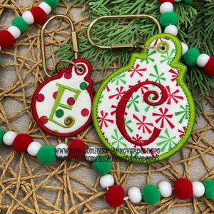 May include: Two round embroidered keychains with gold-tone metal hardware. The smaller keychain features a green letter E on a white background with red and green dots. The larger keychain features a red letter C on a white background with red and green starburst patterns.