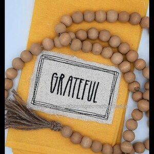 Puede incluir: Un pa&ntilde;o amarillo doblado con un parche de lino que dice "Grateful" en letras negras. Una guirnalda de cuentas de madera y una borla de yute est&aacute;n dispuestas sobre el pa&ntilde;o.
