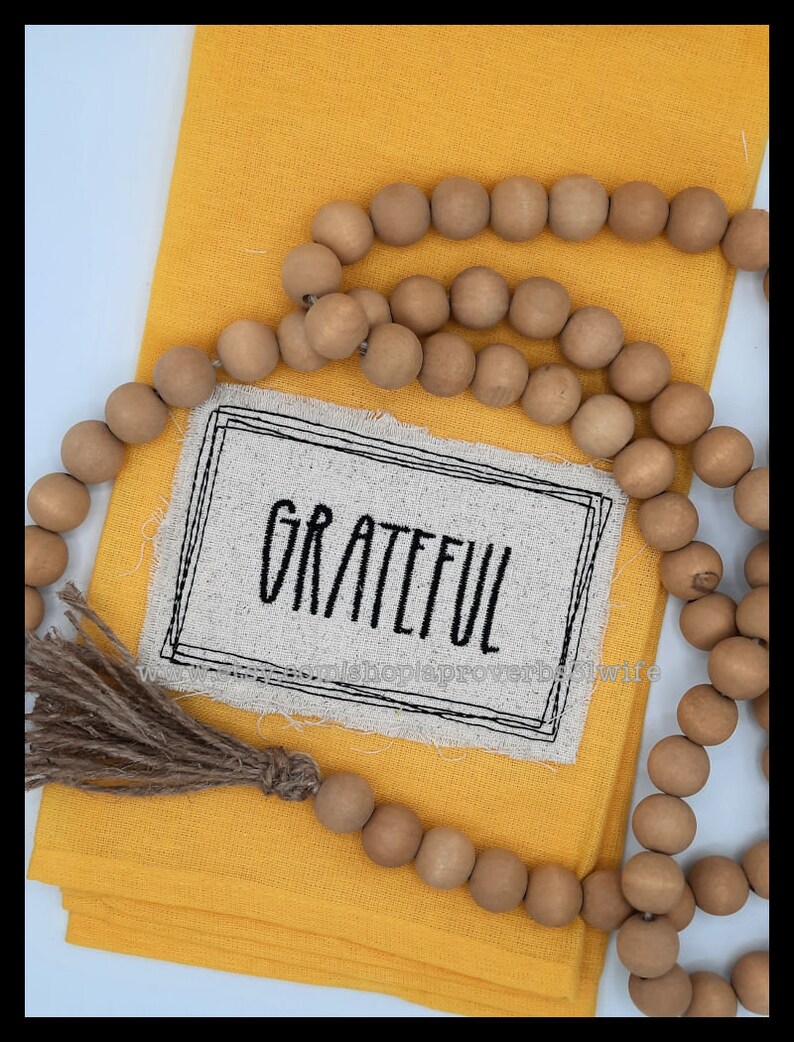 Puede incluir: Un pa&ntilde;o amarillo doblado con un parche de lino que dice "Grateful" en letras negras. Una guirnalda de cuentas de madera y una borla de yute est&aacute;n dispuestas sobre el pa&ntilde;o.