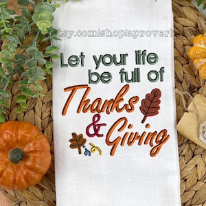 May include: White kitchen towel with the text "Let your life be full of Thanks & Giving" embroidered in orange, red, and green. The text is surrounded by small embroidered leaves and swirls. The towel is on a woven placemat with two small orange pumpkins.