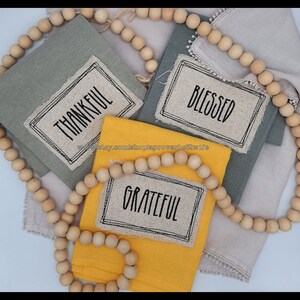 Puede incluir: Servilletas de lino dobladas en tonos grises y amarillos, cada una con una etiqueta con las palabras "THANKFUL", "BLESSED" y "GRATEFUL" en negro. Una guirnalda de cuentas de madera enmarca las servilletas. Ideal para la decoraci&oacute;n del hogar.