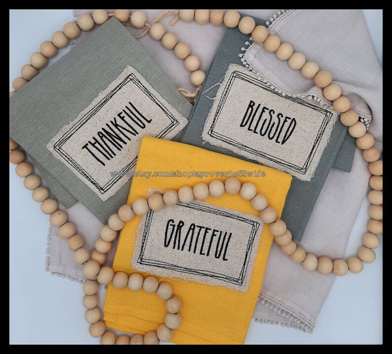 Puede incluir: Servilletas de lino dobladas en tonos grises y amarillos, cada una con una etiqueta con las palabras "THANKFUL", "BLESSED" y "GRATEFUL" en negro. Una guirnalda de cuentas de madera enmarca las servilletas. Ideal para la decoraci&oacute;n del hogar.