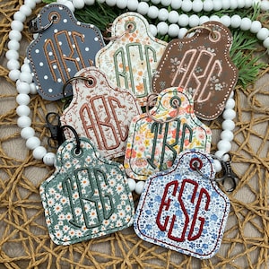 Könnte beinhalten: Eine Sammlung personalisierter Schlüsselanhänger in verschiedenen floralen und gemusterten Designs. Jeder Schlüsselanhänger hat gestickte Initialen in kontrastierenden Farben, mit einem Metallring und Verschluss zur Befestigung. Die Schlüsselanhänger sind in Blau-, Braun- und Grüntönen gehalten.