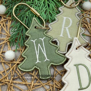 May include: Three Christmas tree-shaped keychains featuring embroidered letters. One is dark green with a white W, one is light sage green with a white R, and one is cream with a dark green D. Each has a gold-toned metal ring and stitched edges.