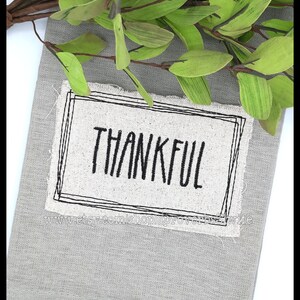 Puede incluir: Un pa&ntilde;o decorativo gris con textura de lino con la palabra "THANKFUL" en escritura negra dentro de un rect&aacute;ngulo con contorno negro. Hojas y ramas artificiales verdes y marrones dispuestas en la parte superior.