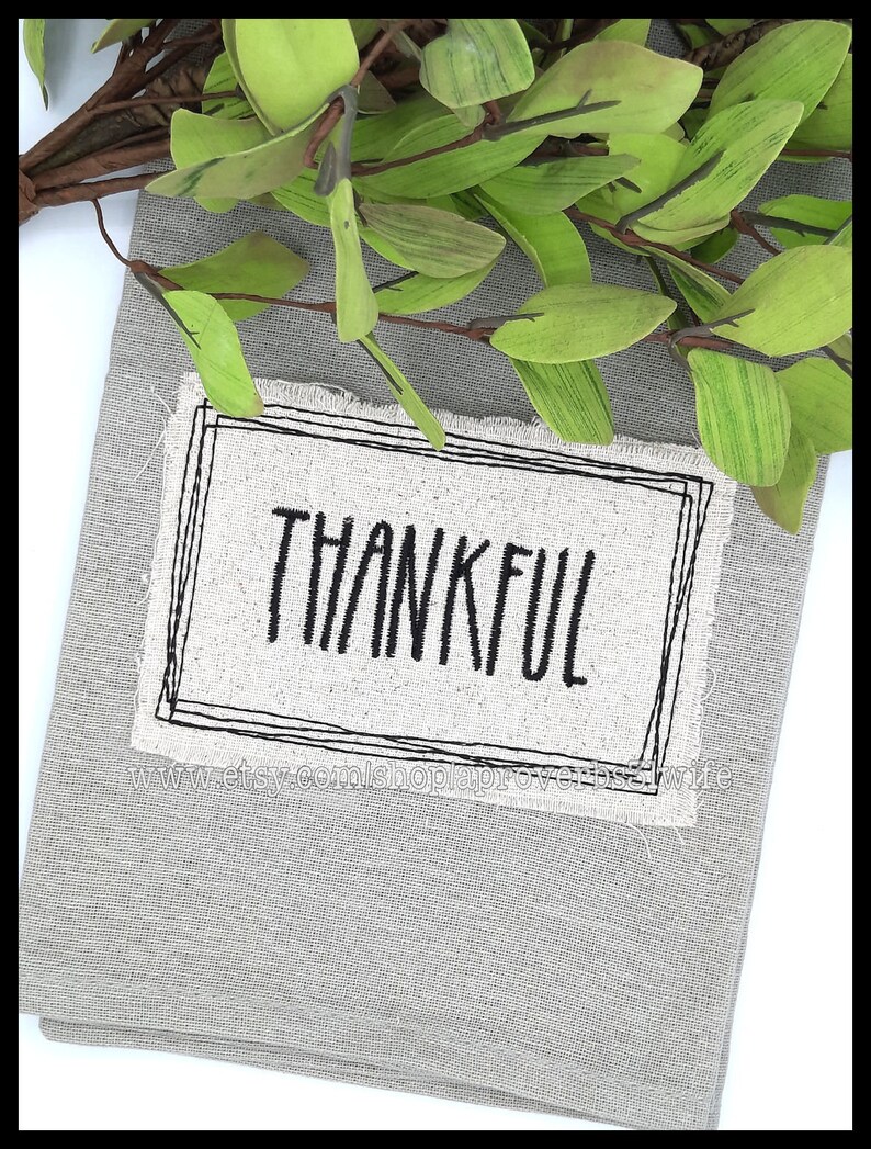 Puede incluir: Un pa&ntilde;o decorativo gris con textura de lino con la palabra "THANKFUL" en escritura negra dentro de un rect&aacute;ngulo con contorno negro. Hojas y ramas artificiales verdes y marrones dispuestas en la parte superior.