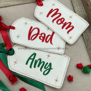 May include: Three white felt gift tags with gold stitching and gold glitter stars. The tags are decorated with embroidered names: "Mom", "Dad", and "Amy".