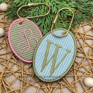 May include: Two oval-shaped fabric keychains with gold-toned metal rings. One keychain features olive green fabric with white stripes and a pink embroidered letter B. The other features light blue fabric with white stripes and an olive green letter W.