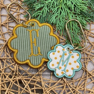 Op de afbeelding: Twee bloemvormige sleutelhangers met goudkleurige metalen sluitingen. De grotere sleutelhanger heeft een groen-wit gestreepte stof met een goud geborduurde letter E. De kleinere heeft een witte stof met gouden stippen en een lichtblauw geborduurde letter M.