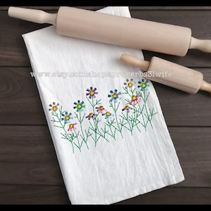 May include: White kitchen towel with embroidered flowers in shades of blue, pink, and yellow. The towel is folded and placed on a wooden surface with two wooden rolling pins.