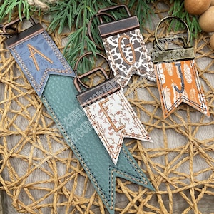 May include: Four personalized keychains with different designs. One is blue with the letter "A", another is white with the letter "L", a third is brown and white with the letter "G", and the last is orange with the letter "J". Each keychain has a metal ring.