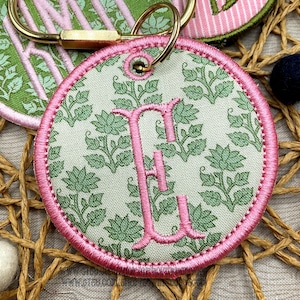 May include: A circular fabric patch featuring a light beige background with a repeating green floral pattern and a large, embroidered pink letter E in the center. The patch has a thick, stitched pink border and a gold-toned metal keyring attached.