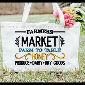May include: White tote bag with the text "Farmers Market Farm to Table Honey Produce Dairy Dry Goods" embroidered in black, blue, and yellow. The bag is sitting on green grass with a bouquet of pink flowers.