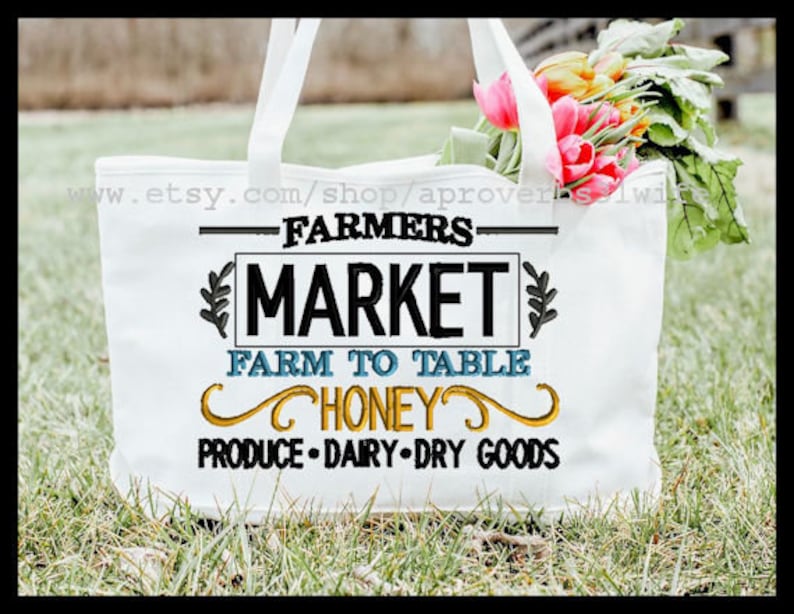 May include: White tote bag with the text "Farmers Market Farm to Table Honey Produce Dairy Dry Goods" embroidered in black, blue, and yellow. The bag is sitting on green grass with a bouquet of pink flowers.