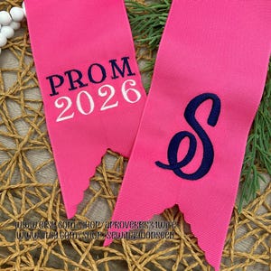 May include: Two bright pink ribbons with a textured surface. One ribbon has the text "PROM 2026" in white, and the other has a navy blue script letter "S". The ribbons have a jagged bottom edge.